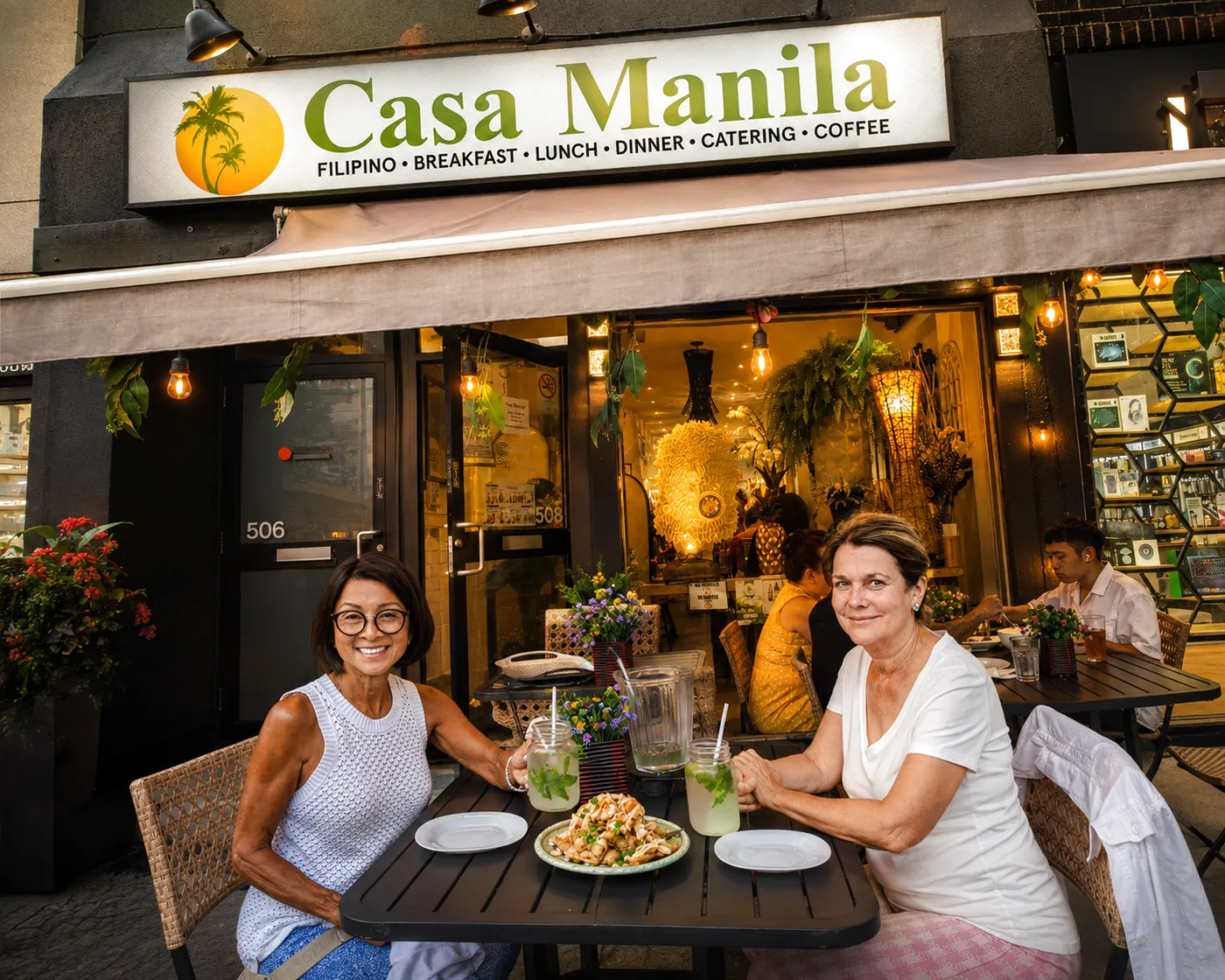 Two diners share sisig and calamansi lemonades on the Casa Manila patio on the Danforth at golden hour, storefront sign glowing warm above them